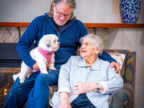 BAPTCARE ABBEY GARDENS resident mother with her son and his dog in the lounge area next to the fireplace smiling