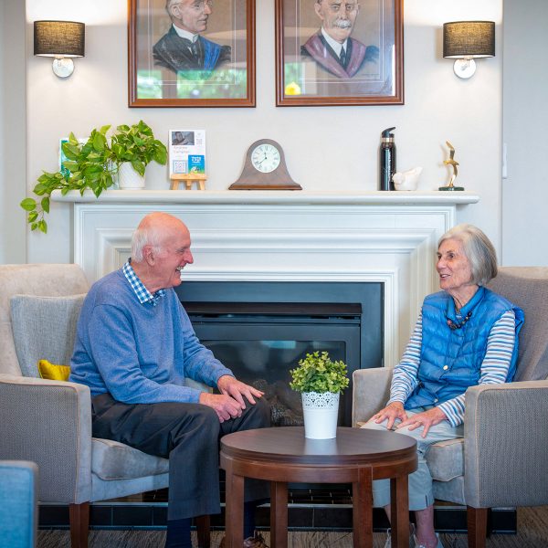 BAPTCARE HEDLEY SUTTON Resident couple having an in depth conversation and laugh in front of the lounge area fireplace