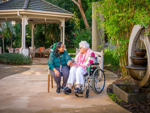 BAPTCARE HEDLEY SUTTON Resident sitting outside in garden courtyard near gazebo and fountain chatting with a staff member