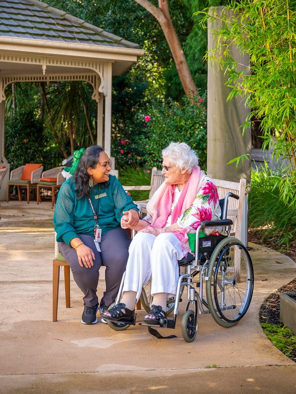 BAPTCARE HEDLEY SUTTON Resident sitting outside in garden courtyard near gazebo and fountain chatting with a staff member
