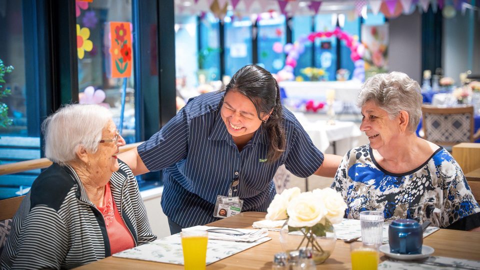 BAPTCARE Wyndham Lodge two resident friends at an event in dining room sharing a laugh with a staff member