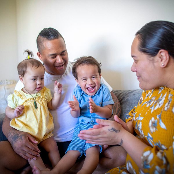 FAMILY COMMUNITY Young mum and dad on couch with their toddler daughter and older toddler brother all smiling together being playful