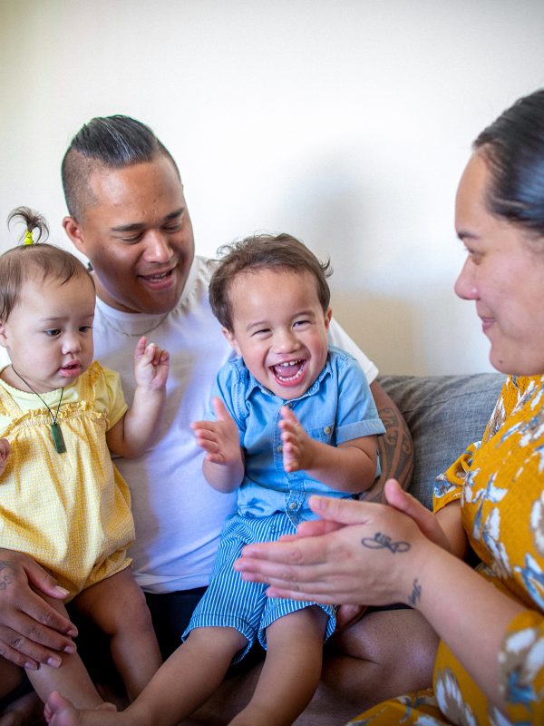 FAMILY COMMUNITY Young mum and dad on couch with their toddler daughter and older toddler brother all smiling together being playful