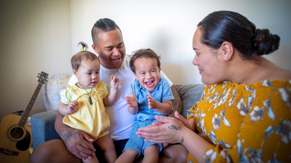 FAMILY COMMUNITY Young mum and dad on couch with their toddler daughter and older toddler brother all smiling together being playful