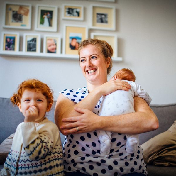 FAMILY COMMUNITY Young mum holding her infant smiling with another child next to her on the couch sucking her thumb