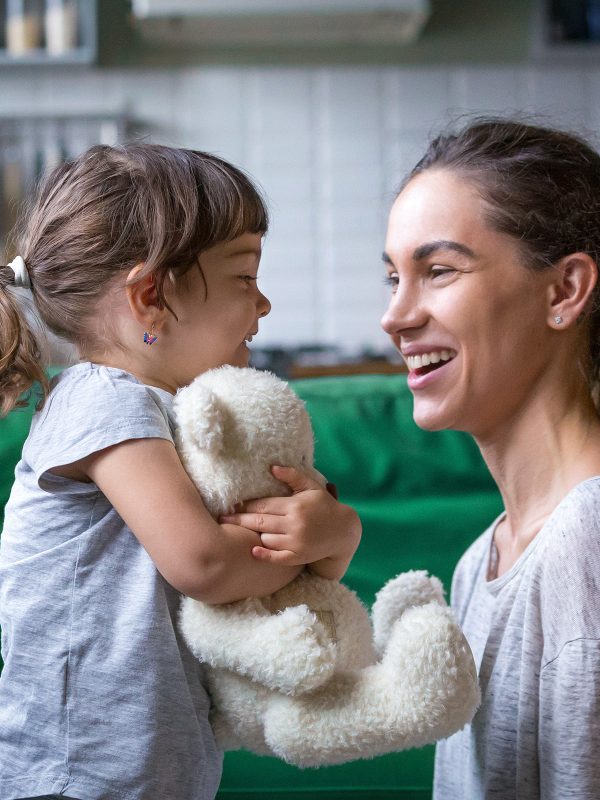 Young single woman foster carer laughing with a young girl in her care who is hugging a teddy bear