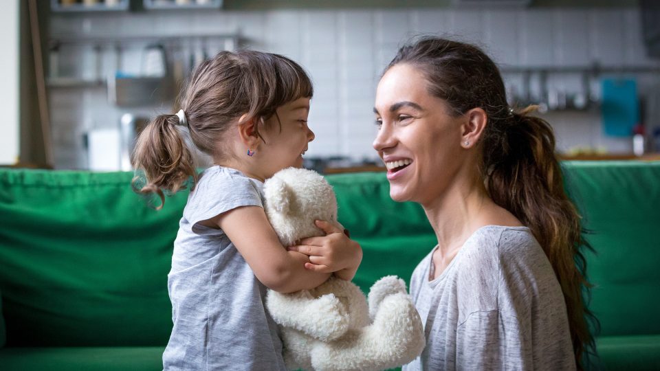 Young single woman foster carer laughing with a young girl in her care who is hugging a teddy bear