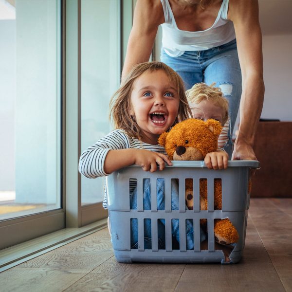 Two young foster care siblings showing pure joy being pushed around the kitchen in a washing basket by a foster carer