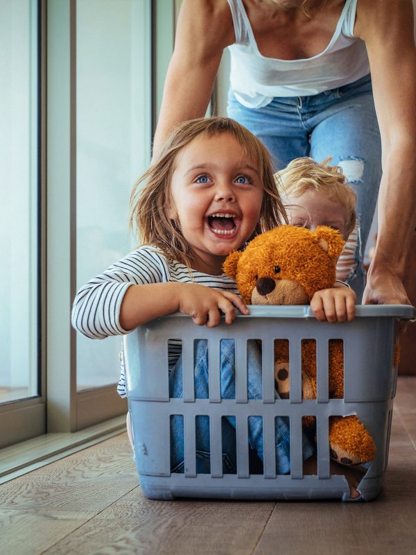 Two young foster care siblings showing pure joy being pushed around the kitchen in a washing basket by a foster carer