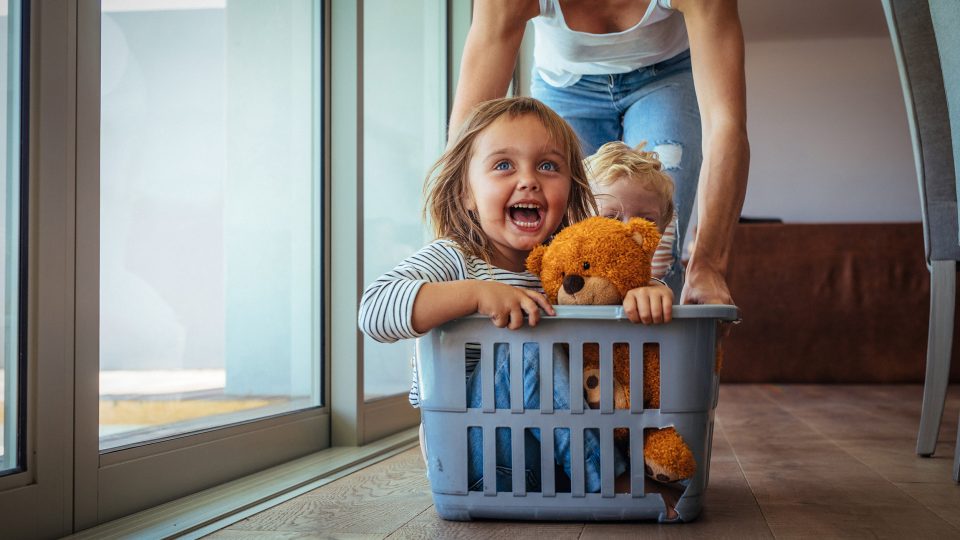 Two young foster care siblings showing pure joy being pushed around the kitchen in a washing basket by a foster carer