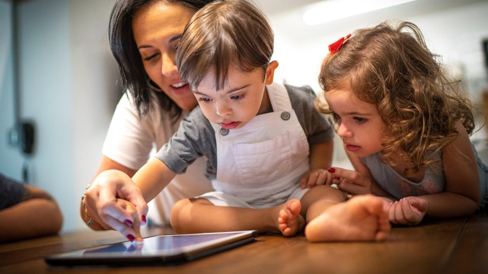 DISABILITY mum teaching two kids, one with Downs syndrome how a digital tablet works, both are concentrating on what she is saying