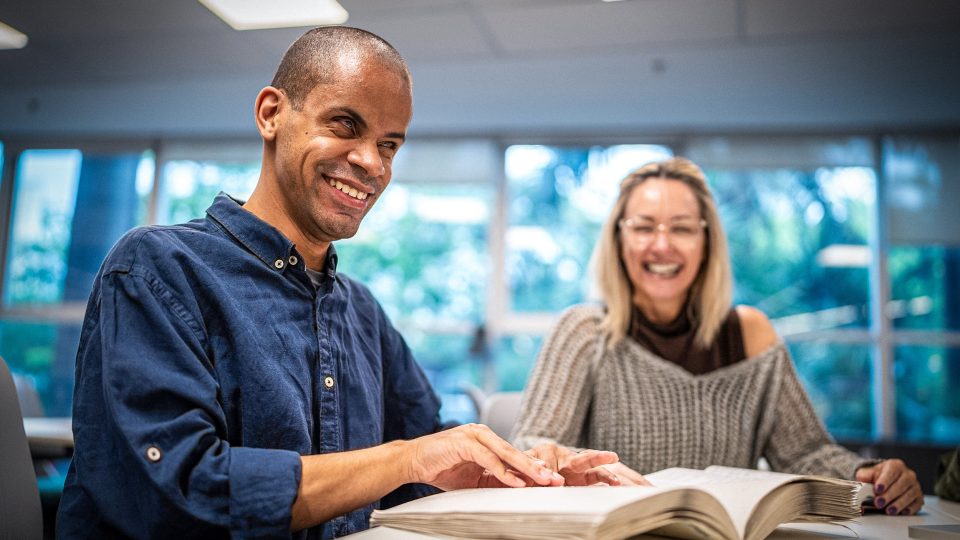 DISABILITY blind young man smiling while reading a braille book next to a female friend