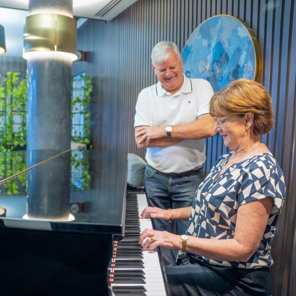 Retirement Living residents inside TheOrchards hub she is playing the piano while her husband stands listening