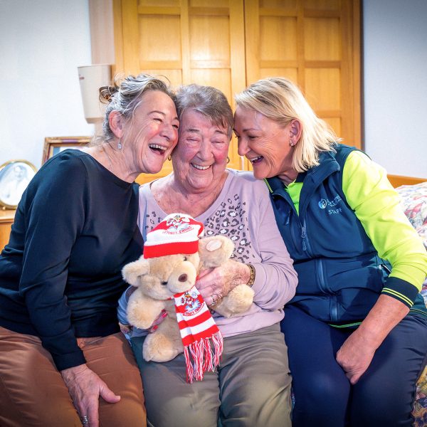 BAPTCARE KARINGAL Resident with both daughters playing around together in her bedroom holding a AFL Sydney Swans teddy bear