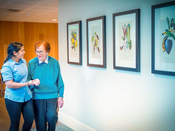 BAPTCARE Wyndham Lodge resident and staff member walking and chatting as they pass a gallery of colourful nature prints on the wall