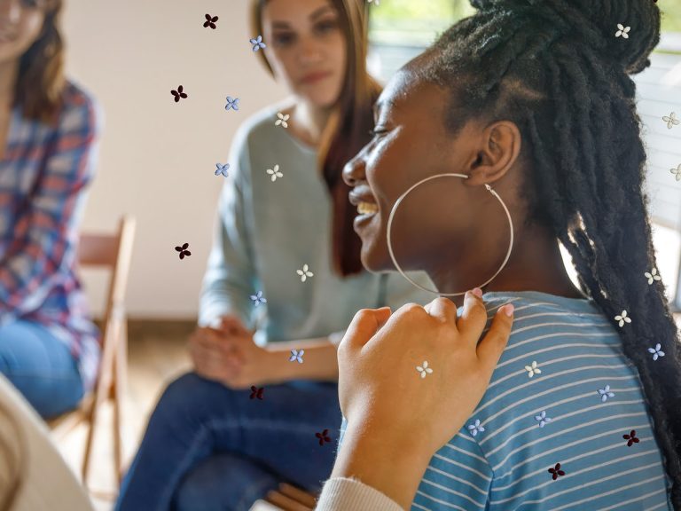 Woman sitting in a circle indoors with a group on wooden fold out chairs, with a hand placed on her shoulder.