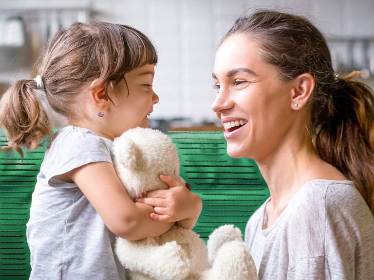 Mother and child smiling at each other, at eye level. Child is holding a white teddy bear.
