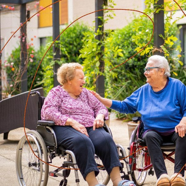 Two residential aged care residents, sitting in wheel chairs, conversing outdoors.