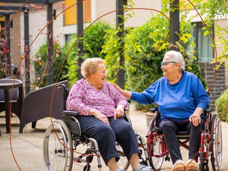 Two residential aged care residents, sitting in wheel chairs, conversing outdoors.